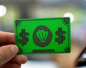 May include: A rectangular green card featuring a dark green border and corner accents. The center displays a dark green circular emblem with a seashell illustration, flanked by two dark green dollar signs on either side of the central icon.