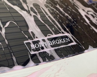 May include: A white sticker with the text "BORN BROKEN" on a black car window covered in white soap suds.