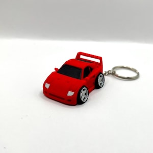 May include: A red toy car keychain with a silver keyring. The miniature car has black windows, white headlights, and silver wheels. The car's design is a stylized representation of a sports car.