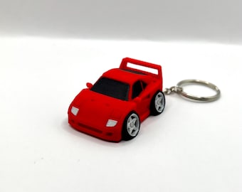 May include: A red toy car keychain with a silver keyring. The miniature car has black windows, white headlights, and silver wheels. The car's design is a stylized representation of a sports car.