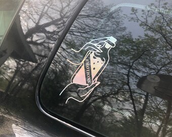 May include: A sticker on a car window with a hand holding a bottle that says "EMOTIONS" on it. The sticker is iridescent and has a rainbow effect.