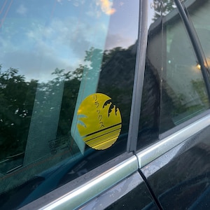 May include: A yellow circular sticker with Japanese text and a palm tree silhouette is attached to a car window. The sticker is on the rear passenger side window of a black car. The background shows a reflection of the sky and trees.