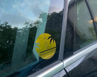 May include: A yellow circular sticker with Japanese text and a palm tree silhouette is affixed to a car window. The sticker is positioned on the rear passenger side window of a black vehicle. The background shows a reflection of the sky and trees.