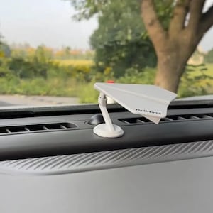 May include: A white airplane-shaped car air freshener with the text "Fly Dreams" on the wing. The air freshener is mounted on a white stand and is placed on a car dashboard. The dashboard is black and has a textured surface.