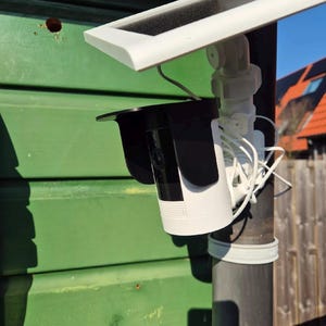 May include: A white and black security camera with a black visor is mounted on a black pole. A white solar panel is attached above the camera. The camera is installed on a green wooden surface.