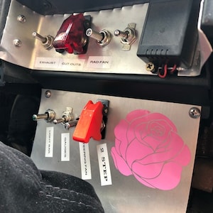 May include: A close-up of a silver panel with various switches and labels. The labels include "Exhaust", "Cut Outs", "Rad Fan", "Light Man", "Gauges", "Steering Fan", and "2 Step". There is a pink rose sticker on the panel.