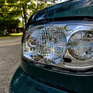 Puede incluir: Primer plano de un faro de coche verde oscuro. El faro tiene un embellecedor cromado y una lente transparente con diseños intrincados. El fondo muestra una calle y árboles borrosos.