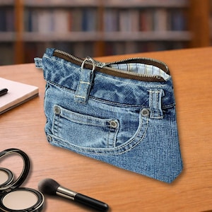 Könnte beinhalten: Eine blaue Jeans-Tasche, die wie eine Hose gestaltet ist, mit einer Vordertasche, Metallnieten, Gürtelschlaufen und einem braunen Reißverschluss. Das Innere ist mit einem hellblau-weiß karierten Stoff ausgekleidet und oben befindet sich ein Metallring.