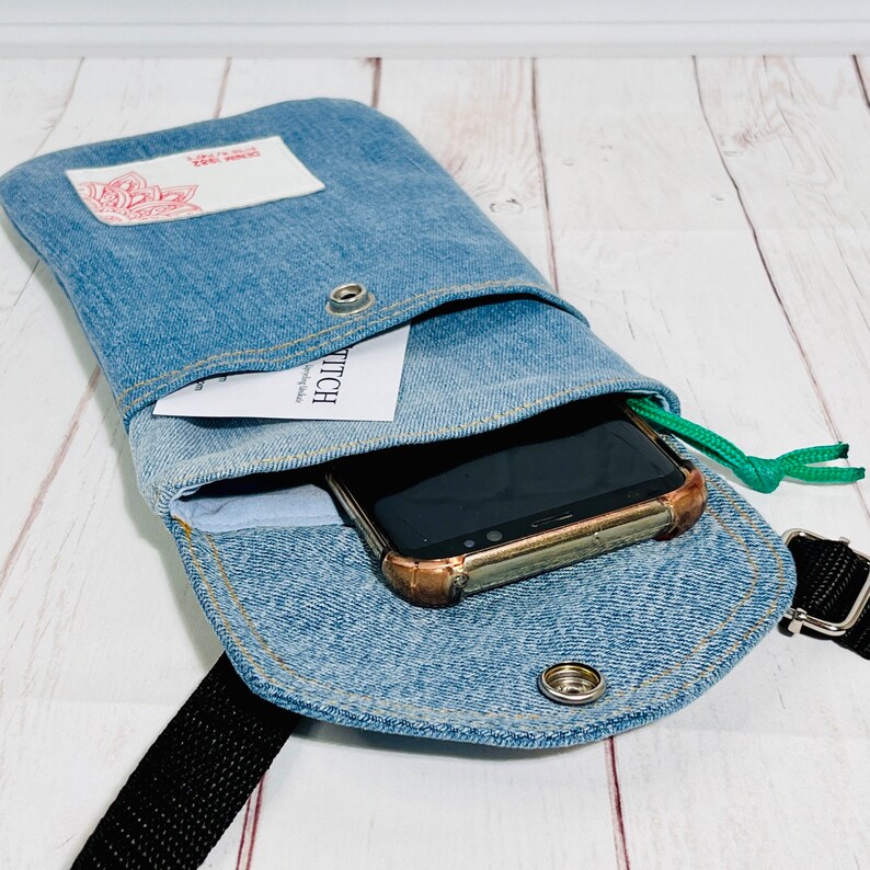 crossbody tasche umhängetasche aus hellblauer upcycling jeans, smartphonetasche zum umhängen, innenansicht liegend und geöffnet im größenvergleich mit einem smartphone