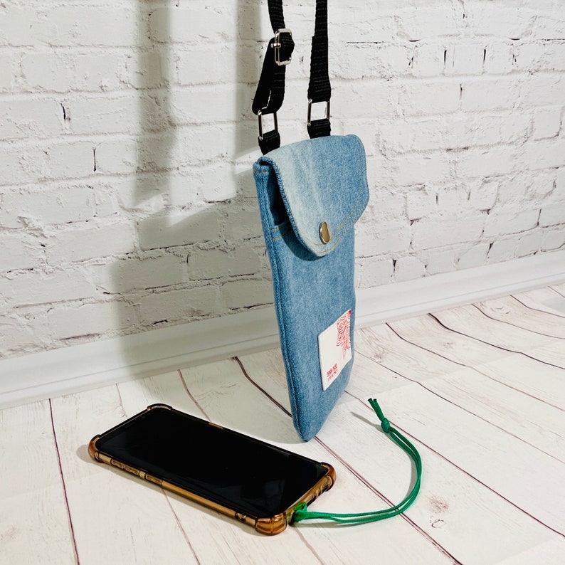 crossbody tasche umhängetasche aus hellblauer upcycling jeans, smartphonetasche zum umhängen, seitenansicht stehend im größenvergleich mit einem smartphone