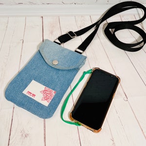 crossbody tasche umhängetasche aus hellblauer upcycling jeans, smartphonetasche zum umhängen, vorderansicht liegend im größenvergleich mit einem smartphone