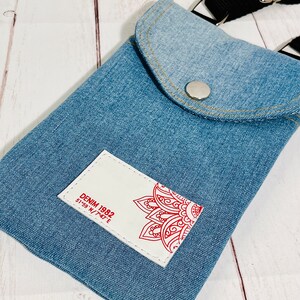 crossbody tasche umhängetasche aus hellblauer upcycling jeans, smartphonetasche zum umhängen, vorderansicht detailansicht weißes logo mit rotem aufdruck