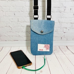crossbody tasche umhängetasche aus hellblauer upcycling jeans, smartphonetasche zum umhängen, vorderansicht stehend im größenvergleich mit einem smartphone