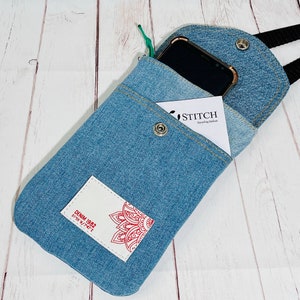 crossbody tasche umhängetasche aus hellblauer upcycling jeans, smartphonetasche zum umhängen, vorderansicht liegend und geöffnet im größenvergleich mit einem smartphone