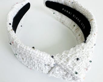 Puede incluir: Diadema de tweed blanco con diseño anudado. La diadema está adornada con pequeñas cuentas de colores. La banda interior es negra y presenta el texto "RONNI BLAKE & CO".
