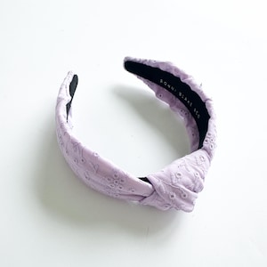 Puede incluir: Una diadema anudada de color lavanda con un delicado patrón bordado. La diadema está hecha de tela suave y tiene un ajuste cómodo. El texto "RONNI BLAKE NYC" está impreso en la parte posterior de la diadema.