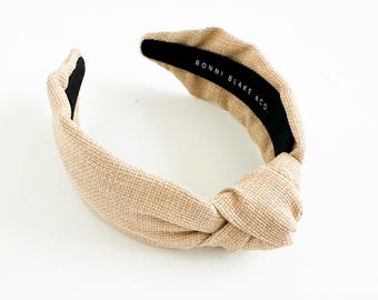 May include: A beige fabric headband with a knotted design. The headband has a textured appearance and a black interior. The text "RONNI BLAKE & CO." is printed on the inside of the headband.
