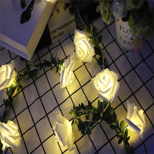 May include: A string of white LED fairy lights with white rose-shaped bulbs and green leaves. The lights are strung together with a clear wire and are lit up.