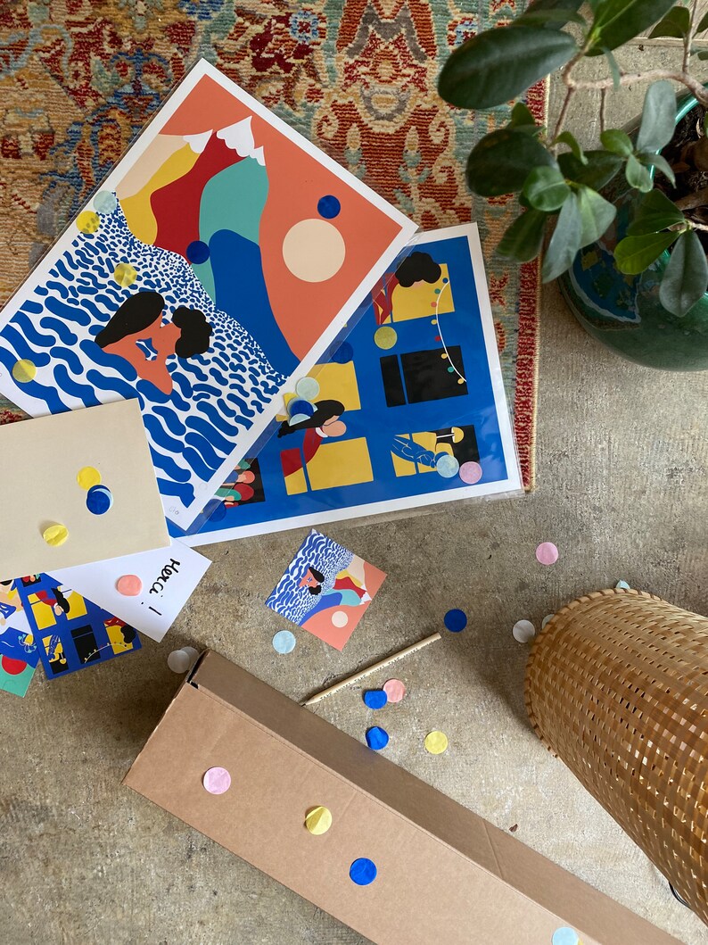 May include: Two rectangular art prints featuring colorful graphic illustrations, one depicting a person swimming in blue waves near mountains and the other showing a building with lit windows, accompanied by a small card with the handwritten text Merci!