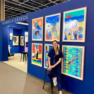 May include: A display wall labeled FRAMES & DESIGN featuring five framed colorful art prints with illustrations of people, landscapes, and a swimming pool. The prints are arranged on a deep blue wall, showcasing various vibrant scenes and graphic styles.