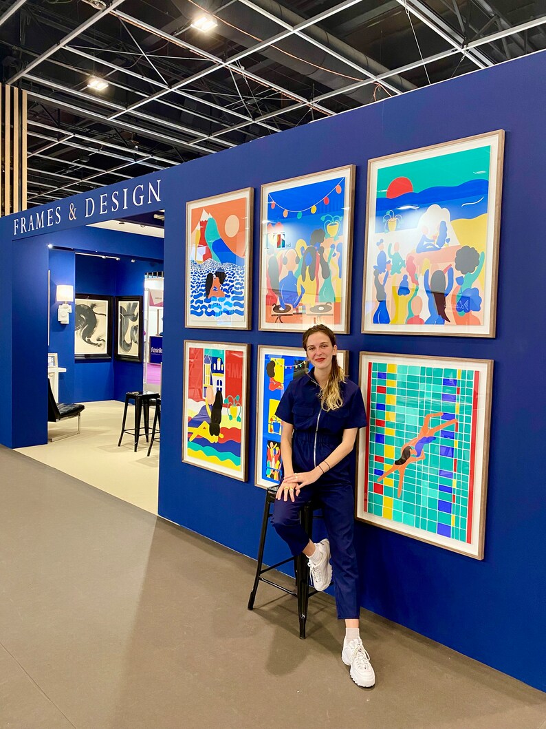 May include: A display wall labeled FRAMES & DESIGN featuring five framed colorful art prints with illustrations of people, landscapes, and a swimming pool. The prints are arranged on a deep blue wall, showcasing various vibrant scenes and graphic styles.
