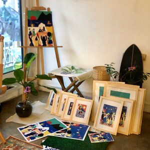 K&ouml;nnte beinhalten: Eine Sammlung gerahmter und ungerahmter Kunstdrucke mit farbenfrohen, stilisierten Illustrationen von Menschen in verschiedenen Umgebungen, die auf einer Staffelei, einem Hocker und dem Boden neben einer Topfpflanze und einem dunklen Objekt stehen.