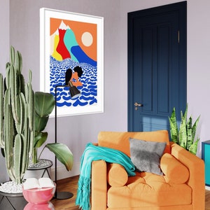 May include: A framed wall art print featuring a stylized illustration of two figures kissing in blue ocean waves beneath a bright orange sky with colorful mountain peaks and a pale sun. The artwork is displayed in a white frame on a light wall.
