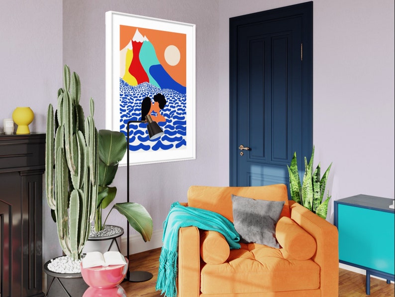May include: A framed wall art print featuring a stylized illustration of two figures kissing in blue ocean waves beneath a bright orange sky with colorful mountain peaks and a pale sun. The artwork is displayed in a white frame on a light wall.