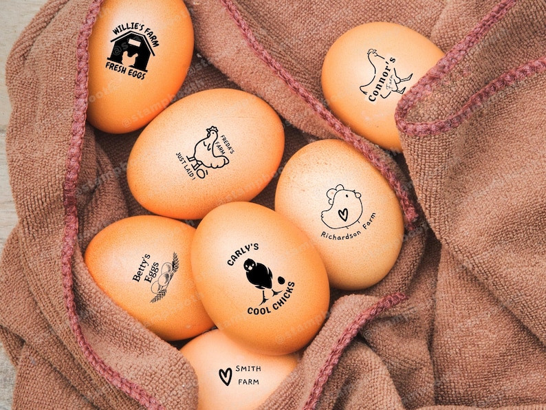 May include: A close-up of brown eggs with various farm-themed stamps. The stamps feature chickens, hearts, and the names of the farms. The eggs are nestled in a brown burlap cloth.