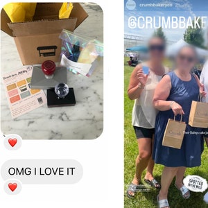 May include: A close-up of a custom rubber stamp with a red handle and a clear acrylic top. The stamp features a small storefront with the text "Crumb Bakery" above it. The stamp is sitting on a gray surface with a white and yellow thank you card and a clear plastic bag with colorful sprinkles.