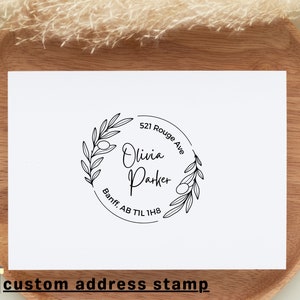 May include: A black and white custom address stamp with an olive branch design. The stamp reads "Olivia Parker" with the address "521 Rouge Ave, Banff, AB T1L 1H8".