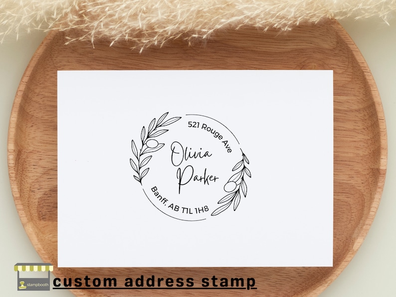May include: A black and white custom address stamp with an olive branch design. The stamp reads "Olivia Parker" with the address "521 Rouge Ave, Banff, AB T1L 1H8".