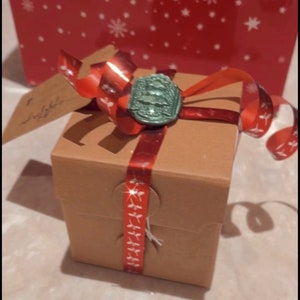 Op de afbeelding: Een kleine bruine cadeaudoos met een rood lint en een groene waszegel. De doos staat op een wit oppervlak.