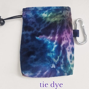 May include: A small rectangular pouch featuring a tie-dye pattern in shades of blue, purple, and teal. The pouch includes a black drawstring closure with a plastic toggle and a silver-coloured metal carabiner attached to the side for easy carrying.
