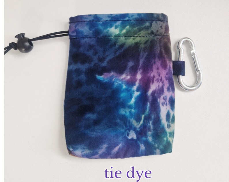 May include: A small rectangular pouch featuring a tie-dye pattern in shades of blue, purple, and teal. The pouch includes a black drawstring closure with a plastic toggle and a silver-coloured metal carabiner attached to the side for easy carrying.