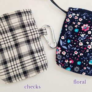 May include: Two small rectangular pouches with drawstring closures and silver carabiner clips. One pouch features a black and white plaid pattern labelled checks, while the other has a dark blue background with a pink, white, and teal floral print labelled floral.