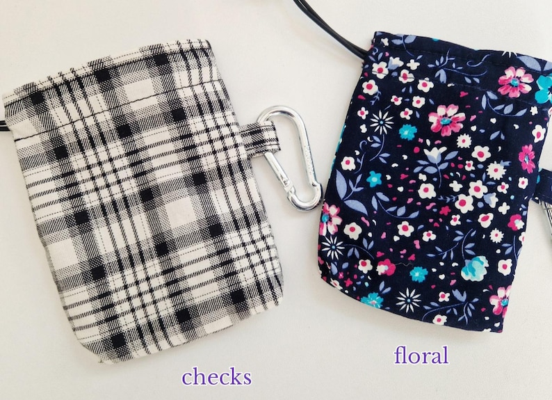 May include: Two small rectangular pouches with drawstring closures and silver carabiner clips. One pouch features a black and white plaid pattern labelled checks, while the other has a dark blue background with a pink, white, and teal floral print labelled floral.