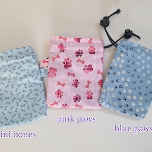 May include: Three rectangular fabric pouches with drawstring closures. The left pouch is light blue with a mini bones pattern. The centre pouch is pink with a pink paws pattern and labelled pink paws. The right pouch is blue with a blue paws pattern and labelled blue paws.