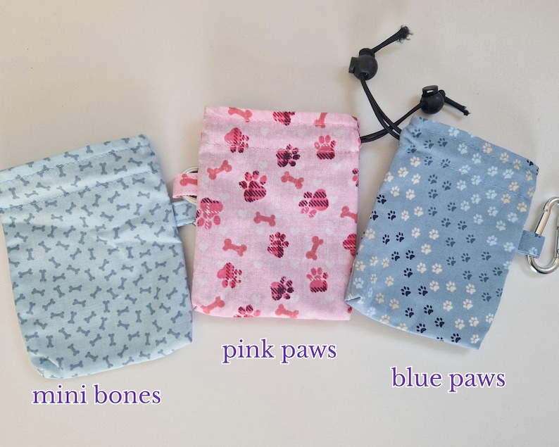 May include: Three rectangular fabric pouches with drawstring closures. The left pouch is light blue with a mini bones pattern. The centre pouch is pink with a pink paws pattern and labelled pink paws. The right pouch is blue with a blue paws pattern and labelled blue paws.