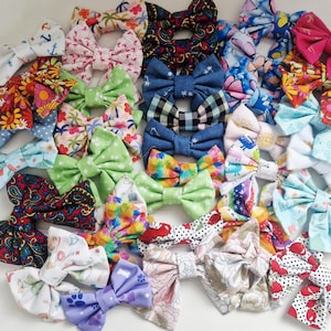 May include: A selection of fabric bows in a variety of patterns and colours. The bows feature floral, paisley, and polka dot designs in shades of blue, green, pink, and red. Some bows have anchor and paw print patterns.