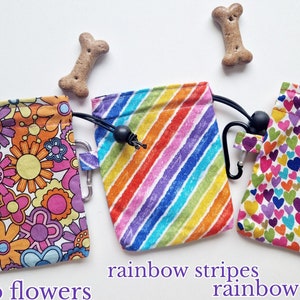 May include: Three fabric treat pouches with drawstring closures and black carabiners. The left pouch features a multi-coloured floral pattern, the centre pouch has diagonal rainbow stripes, and the right pouch displays a pattern of small rainbow hearts.