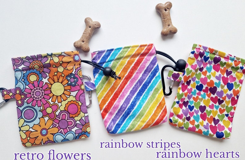 May include: Three fabric treat pouches with drawstring closures and black carabiners. The left pouch features a multi-coloured floral pattern, the centre pouch has diagonal rainbow stripes, and the right pouch displays a pattern of small rainbow hearts.