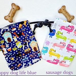 May include: Two fabric treat pouches with drawstring closures and carabiners. The left pouch is dark blue with a pattern of dogs, houses, and pet supplies. The right pouch is white with a pattern of colourful dachshunds. Two bone-shaped treats are included.