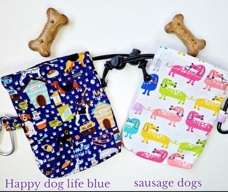 May include: Two fabric treat pouches with drawstring closures and carabiners. The left pouch is dark blue with a pattern of dogs, houses, and pet supplies. The right pouch is white with a pattern of colourful dachshunds. Two bone-shaped treats are included.