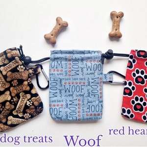 May include: Three fabric treat pouches with drawstrings and carabiners. The left pouch is black with tan bone patterns and the text "GOOD DOG". The centre pouch is light blue with "WOOF" and "bark" text. The right pouch is red with black paw prints.