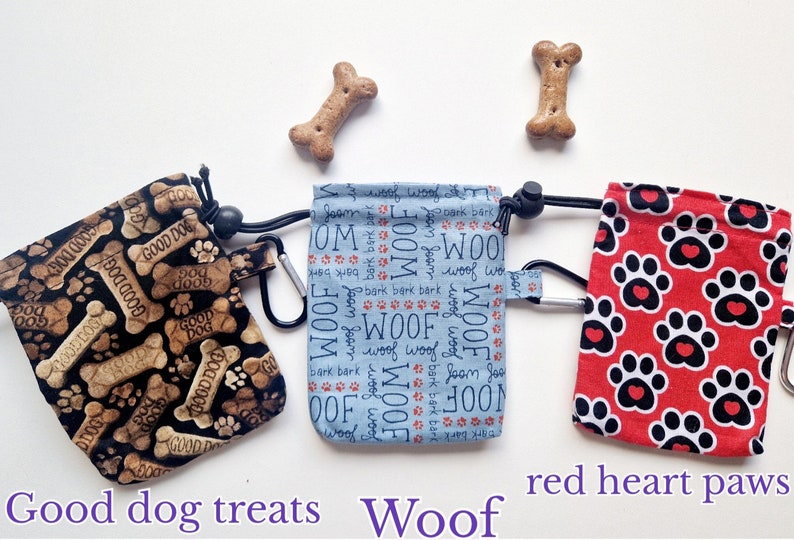 May include: Three fabric treat pouches with drawstrings and carabiners. The left pouch is black with tan bone patterns and the text "GOOD DOG". The centre pouch is light blue with "WOOF" and "bark" text. The right pouch is red with black paw prints.