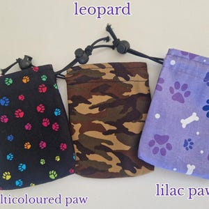 May include: Three rectangular drawstring pouches with metal carabiner clips. The left pouch is black with multicoloured paw prints, the centre pouch has a brown and green camouflage pattern, and the right pouch is lilac with dark purple paw prints and white bones.