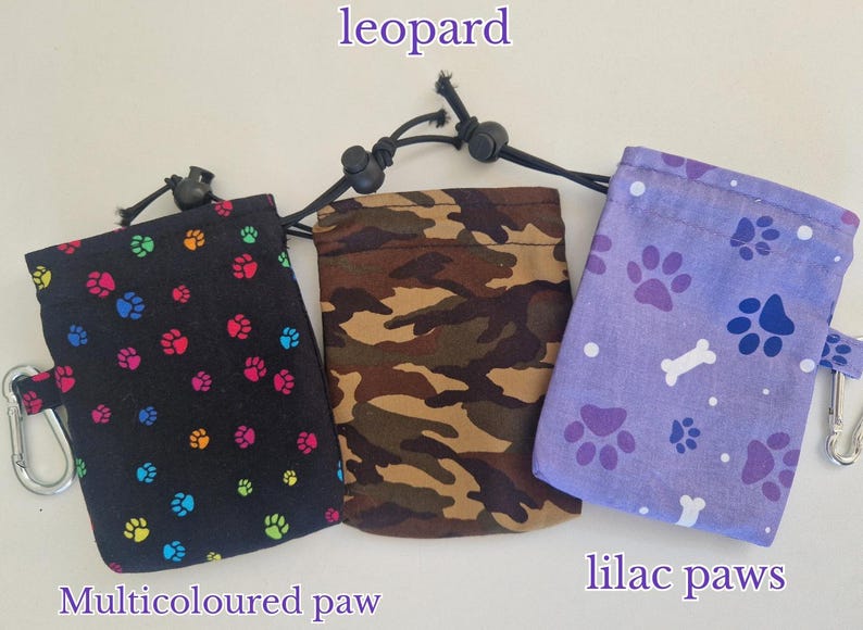 May include: Three rectangular drawstring pouches with metal carabiner clips. The left pouch is black with multicoloured paw prints, the centre pouch has a brown and green camouflage pattern, and the right pouch is lilac with dark purple paw prints and white bones.