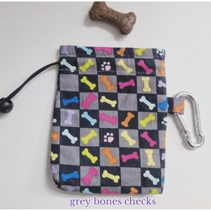 May include: A rectangular drawstring pouch featuring a black and grey checkered pattern with colourful dog bones and paw prints. The pouch includes a black drawstring cord with a plastic toggle and a silver metal carabiner attached to the side.