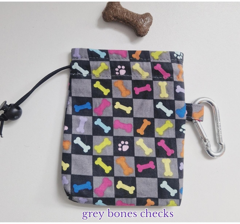 May include: A rectangular drawstring pouch featuring a black and grey checkered pattern with colourful dog bones and paw prints. The pouch includes a black drawstring cord with a plastic toggle and a silver metal carabiner attached to the side.